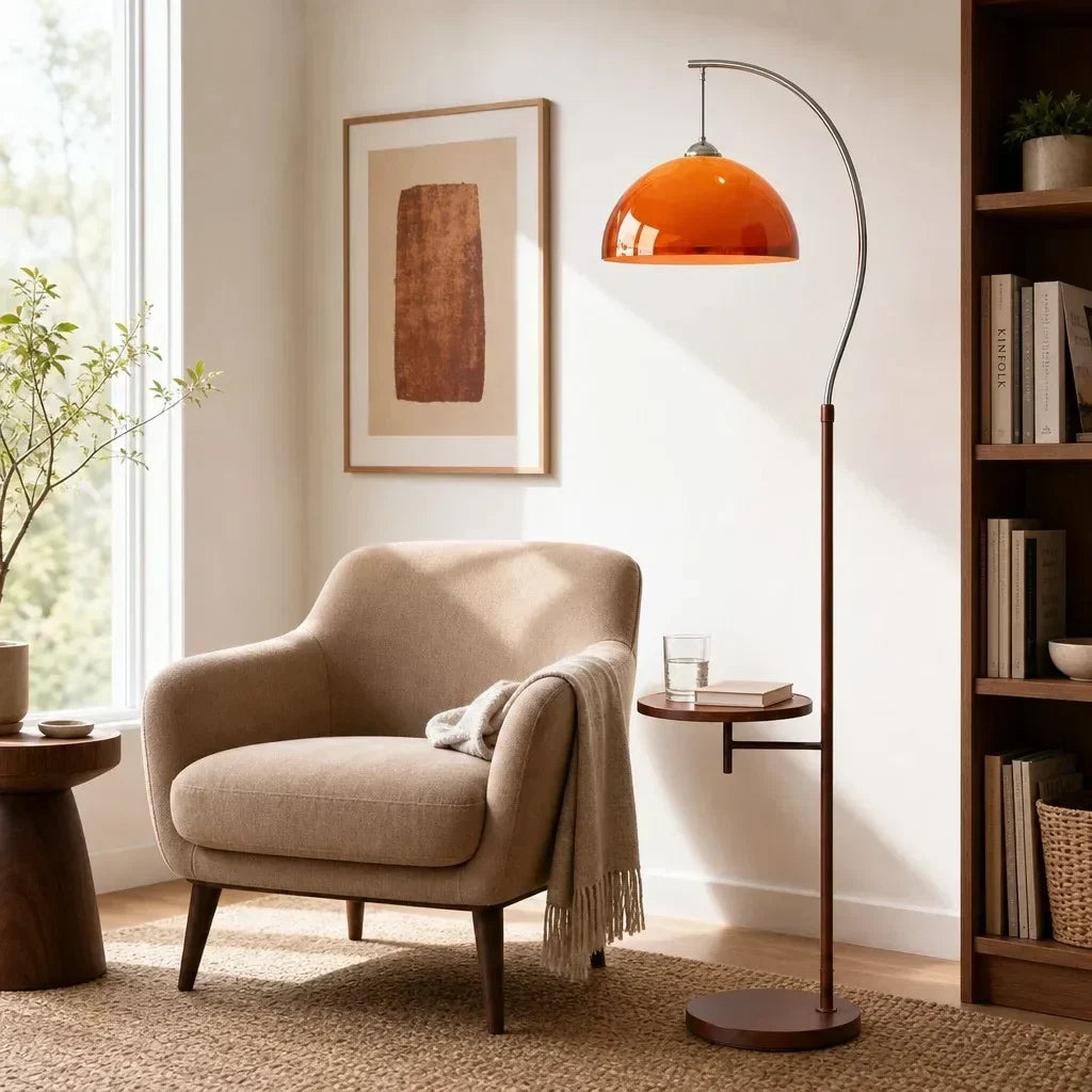 An orange dome floor lamp with a curved dark metal stand and round base, placed beside a beige armchair in a styled living room with a window, framed art, and a bookshelf.