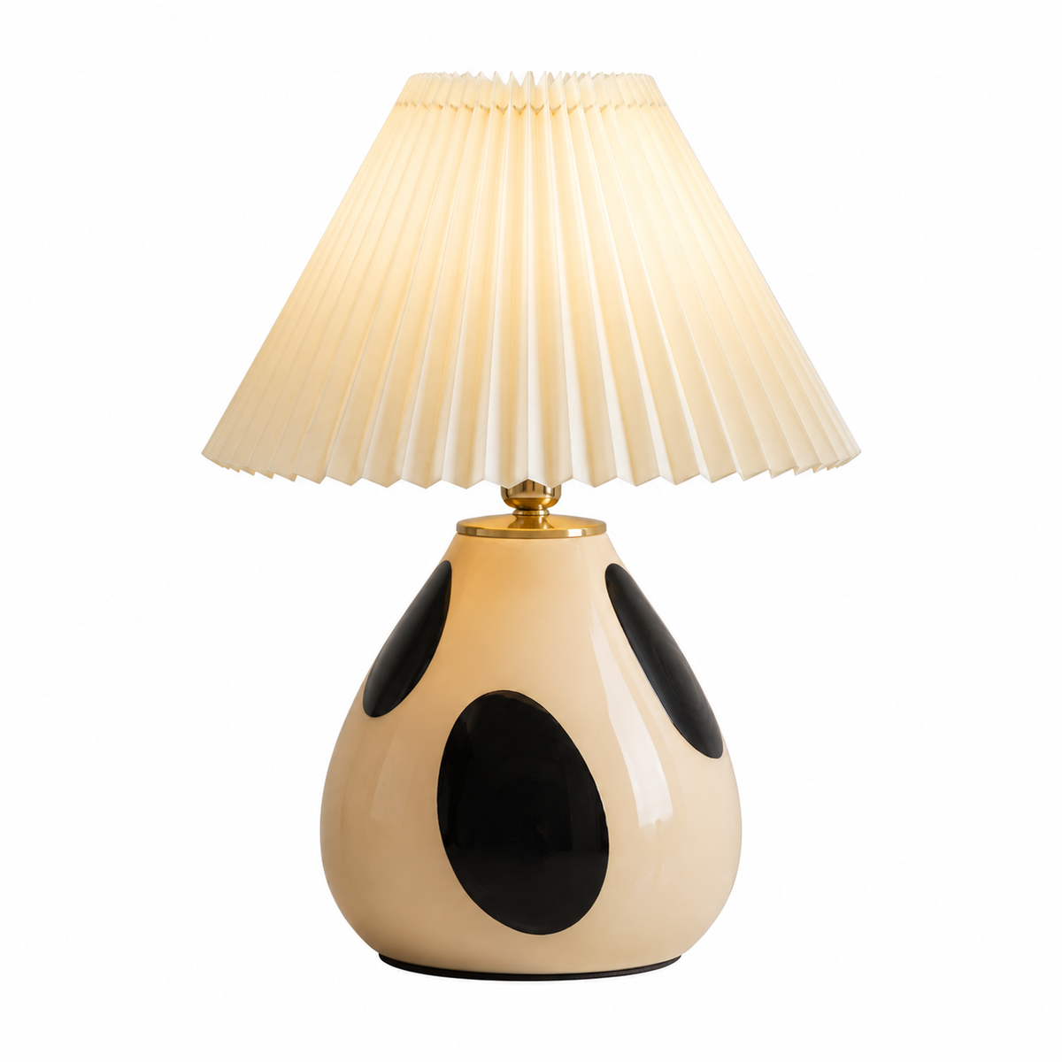A table lamp with a pleated cream shade and a glossy cream ceramic base decorated with black spots, illuminated with a warm glow.