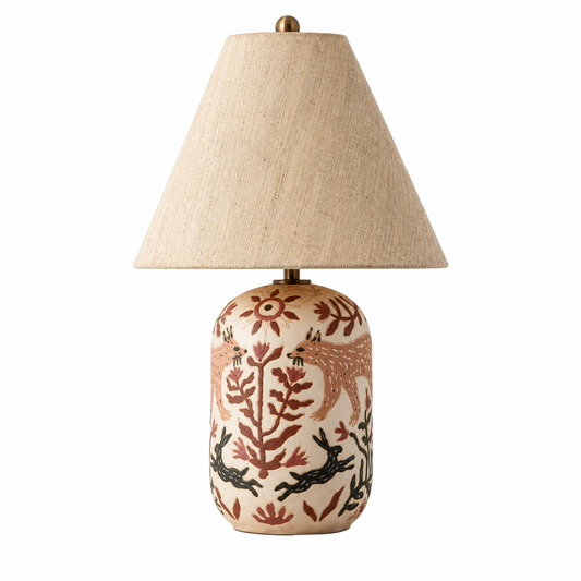 A table lamp with a textured beige fabric shade and a cylindrical ceramic base decorated with hand-painted Native American style motifs including foxes, deer, and floral patterns in earthy red, brown, and black on a cream background.